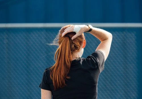 Rear view of a red-haired woman wearing a black t-shirt with her hand on her head in front of a blue fence