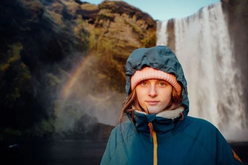 Young woman in a blue jacket with a pink hat stands in front of a waterfall landscape with a rainbow, creating a vibrant nature scene.