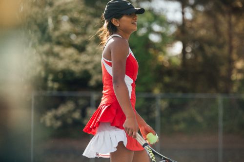 A woman in red tennis dress and black cap holds a tennis ball and racquet on an outdoor court, focusing on a sunlit profile view.
