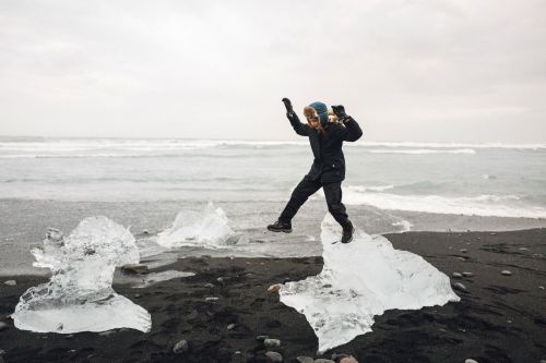 Woman jumping on icebergs on a black sand beach in Iceland, wearing a dark coat, hat, and gloves, with ocean waves in background.