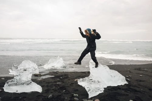 Woman jumping on icebergs on a black sand beach in Iceland, wearing a dark coat, hat, and gloves, with ocean waves in background.