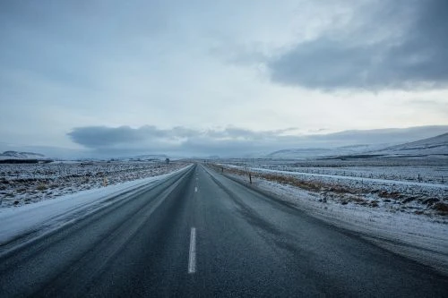 Road stretches into horizon under cloudy sky, flanked by snow-covered fields in a desolate winter landscape. The asphalt is wet and dark.