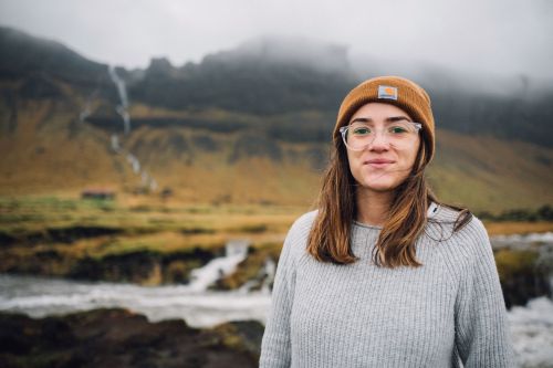 Woman wearing an orange beanie and clear glasses standing in front of a misty mountain landscape and waterfall.