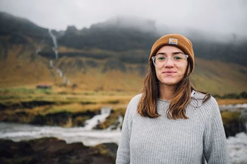 Woman wearing an orange beanie and clear glasses standing in front of a misty mountain landscape and waterfall.