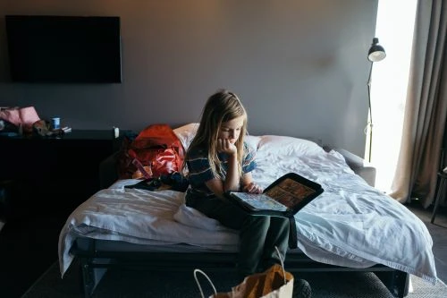Young child sits on a white bed in a grey room and looks at a digital tablet in their lap. An orange backpack sits behind them.