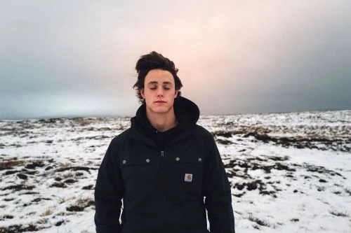 Young man with dark hair in a black winter jacket standing against a snowy landscape under an overcast sky.