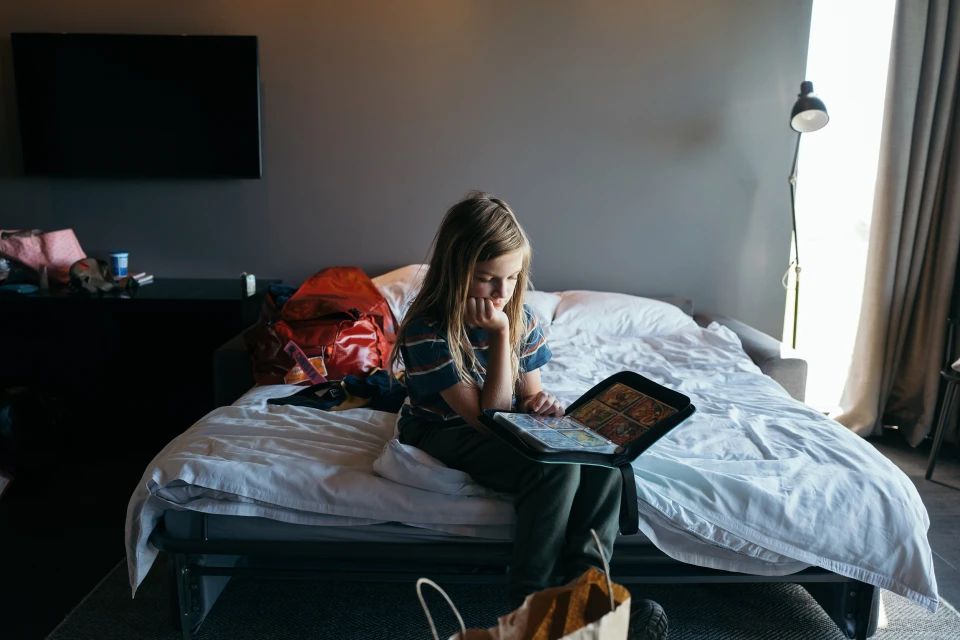 Blond child sits on a bed looking at cards in a binder, with a red backpack and TV in the background in a brightly lit indoor setting.