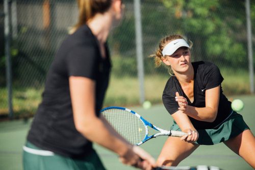 Two women are playing tennis on an outdoor court in daylight. One player focuses on the game while holding her racket.