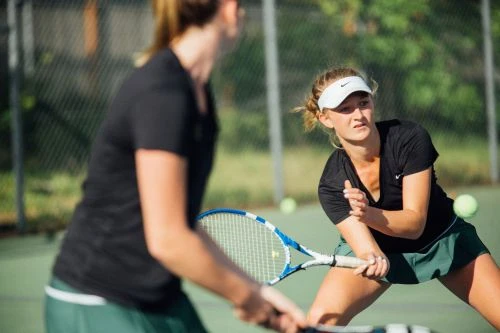 Two women are playing tennis on an outdoor court in daylight. One player focuses on the game while holding her racket. 