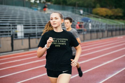 Young woman sprints on an outdoor track, with another runner and stadium seating visible in the background on an overcast day.