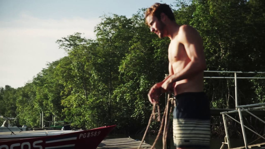 Shirtless man stands on a wooden dock holding rope; a boat is visible in the foreground and a forest makes up the background.