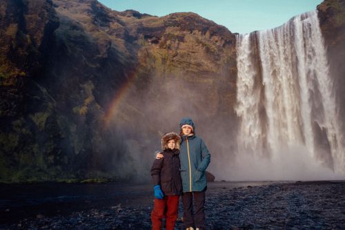 Two children in winter coats stand before Skógafoss Waterfall in Iceland, a rainbow arches over the green mountain landscape.