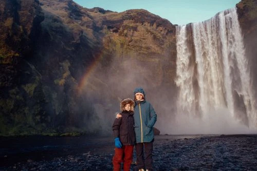 Two children in winter coats stand before Skógafoss Waterfall in Iceland, a rainbow arches over the green mountain landscape.