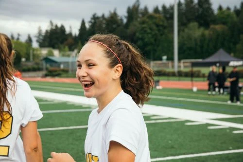 Smiling young woman with a ponytail and headband on a sports field, with another athlete nearby, and spectators in the background.