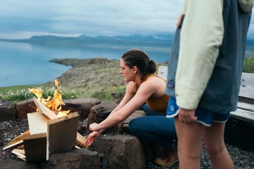 Woman warming her hands by a campfire outdoors with a lake and hills in the background