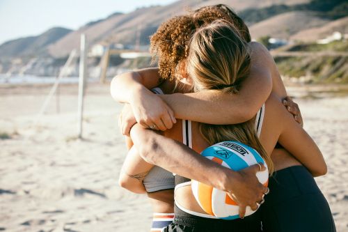 Three young adult women embrace on a beach near mountains, holding a volleyball, wearing athletic wear in bright, natural light.