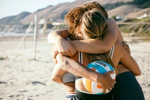 Three young adult women embrace on a beach near mountains, holding a volleyball, wearing athletic wear in bright, natural light.