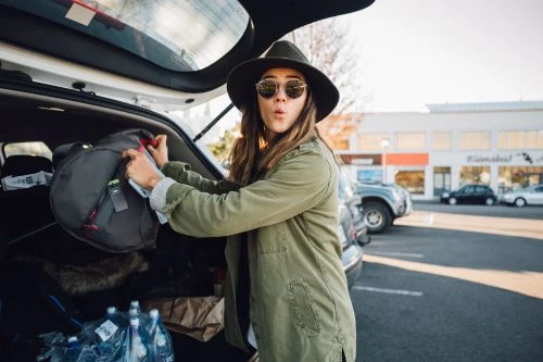Woman in hat and sunglasses loads gray backpack into car trunk outdoors in parking lot near shops on a sunny day.