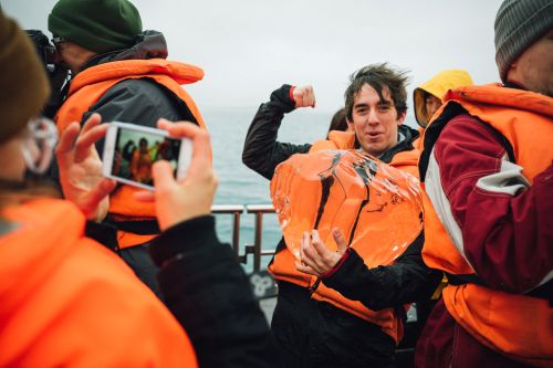 Person holds a life vest on a boat deck with people wearing life vests, cloudy sky, and ocean water in the background.