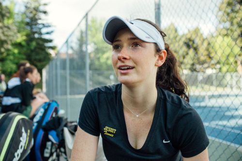 A young woman in a black athletic shirt and visor is looking off-camera, with people and tennis courts visible in the background.