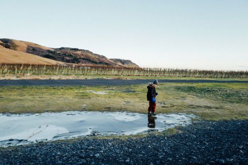A child in a jacket stands on a frozen puddle in a rural landscape with hills and a clear sky in the background on a sunny day.