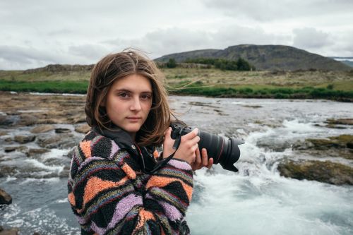 Young woman holds professional camera outdoors with blurred river water and lush green hills in the background on a cloudy day.