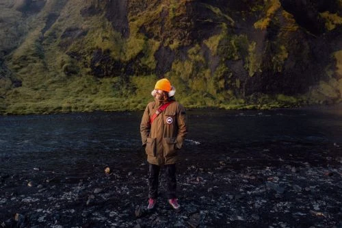Man in a parka coat and bright orange hat stands on a rocky shore near a dark river and a mossy green rockface in Iceland.