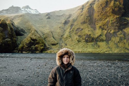 A child stands in a hooded winter coat in a rocky landscape near a river, with mountains covered in green moss in the background.
