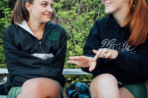 Two young women sitting together outdoors, one wearing a black windbreaker and green skirt and the other a black hoodie