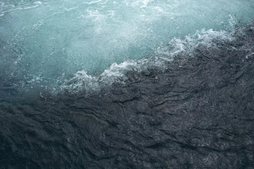 Aerial view of water with dark blue currents, white foam, and light blue accents. Ripples and textures create a dynamic surface.