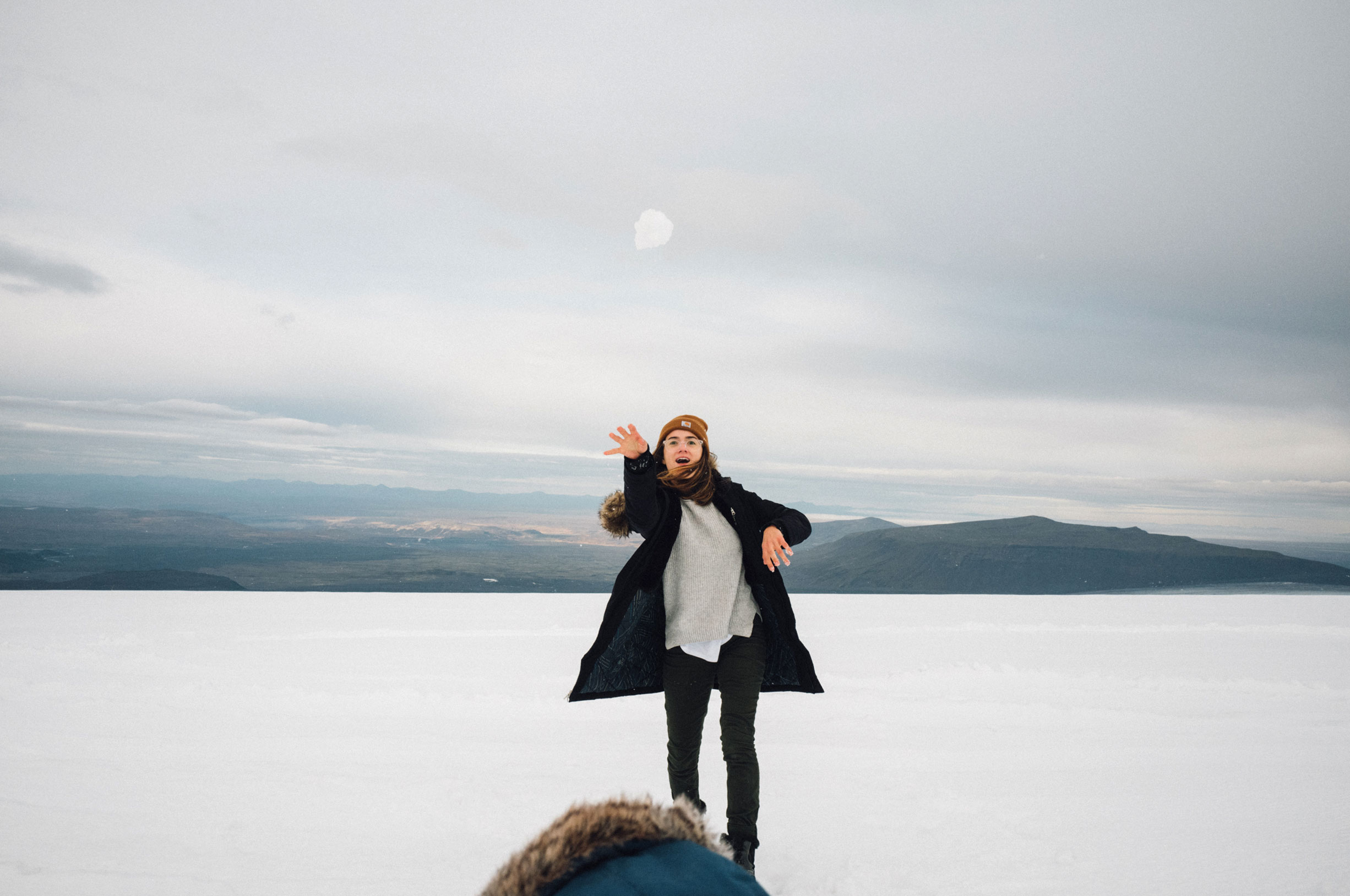 Woman in black coat throws snowball in a snowy landscape with distant mountains under an overcast sky, showcasing winter outdoor activity.