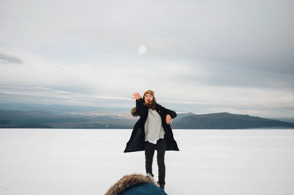 Woman in black coat throws snowball in a snowy landscape with distant mountains under an overcast sky, showcasing winter outdoor activity.