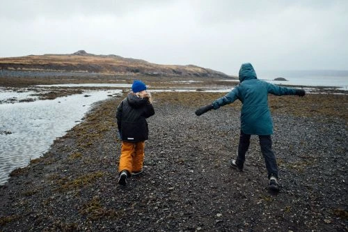 Two children in winter jackets walking along a rocky coastal shore under an overcast sky by Christa Renee