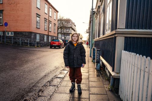 Young girl in a winter coat stands on a sidewalk in a city, with colorful buildings and a red car in the background on a cloudy day.