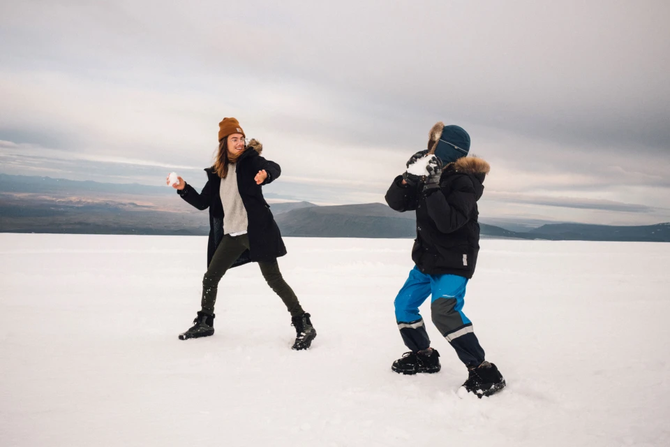 Woman and child having a playful snowball fight in a snowy, open field with a mountain range visible in the distant background.
