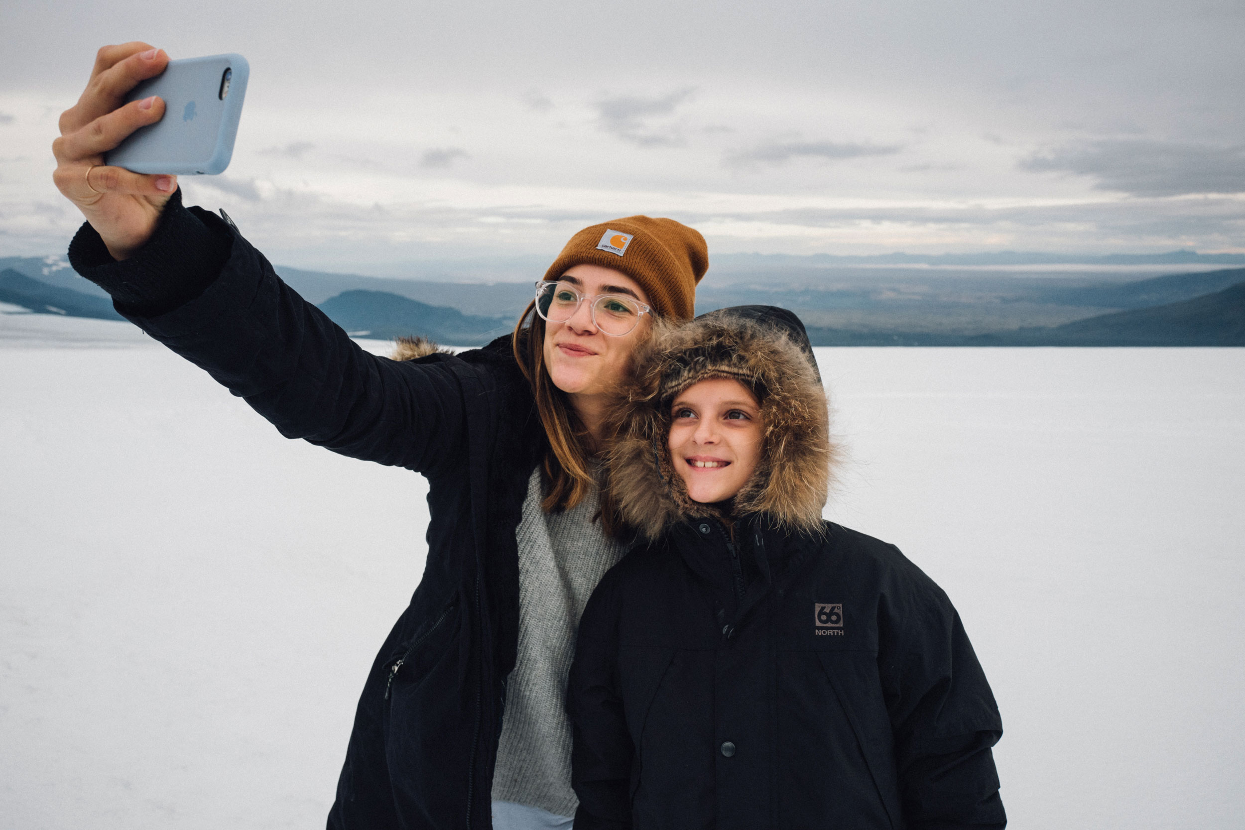 Woman and child take a selfie against a snowy backdrop with distant mountains under a cloudy sky, capturing a family moment outdoors.
