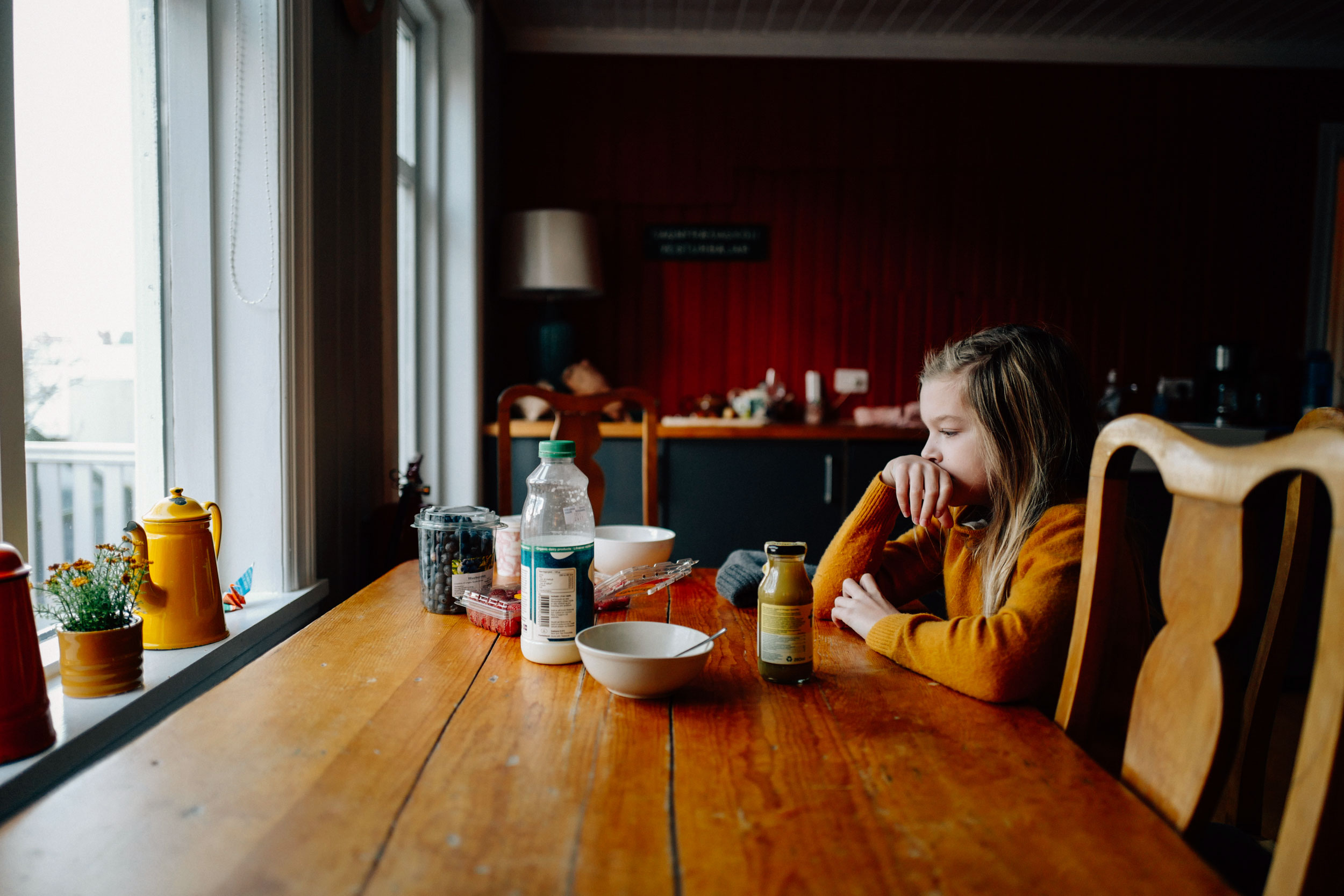 Young girl in a yellow sweater seated at a wooden dining table near a window by photographer Christa Renee