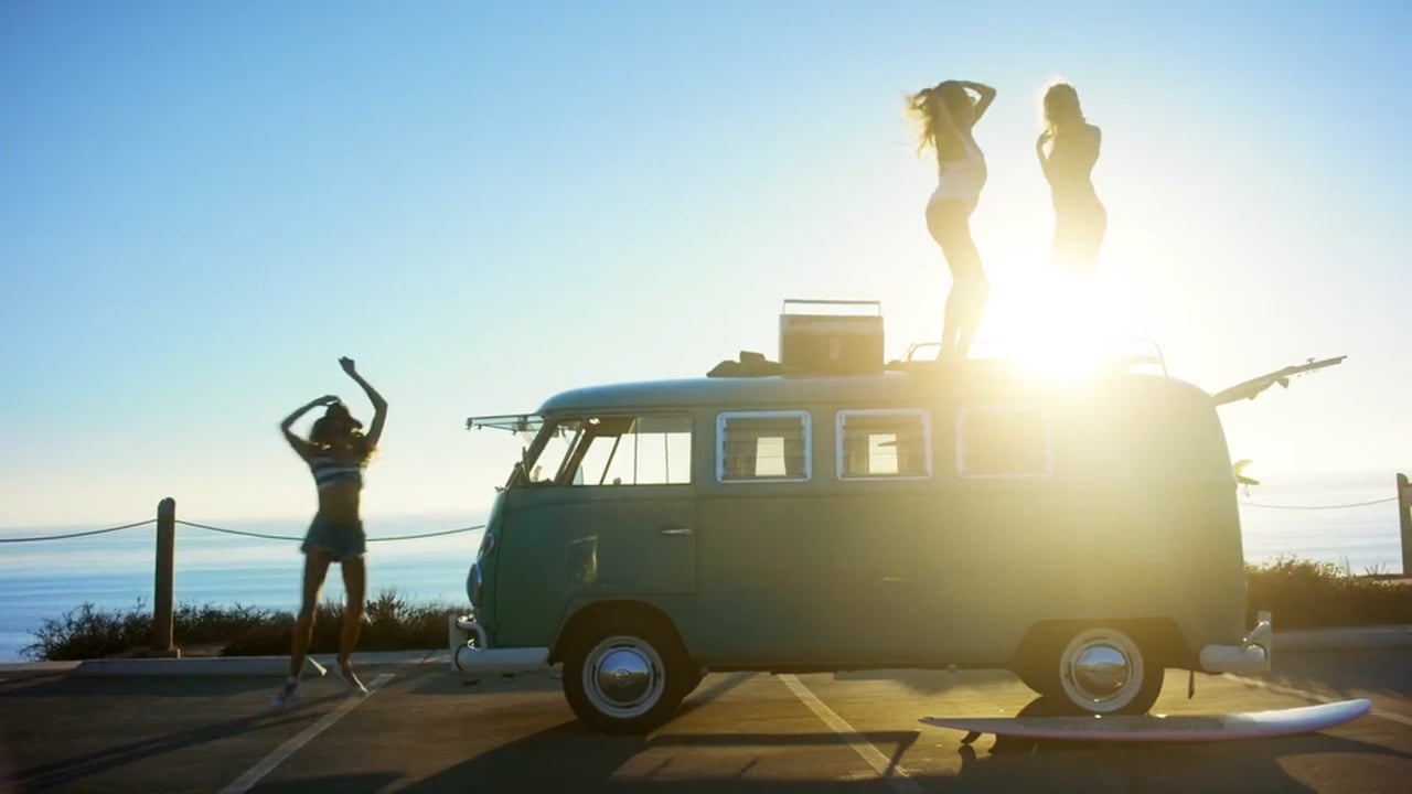 Three women dancing on a vintage van by the ocean in bright sunlight; light teal van with surfboards, blue sky, and sparkling water.