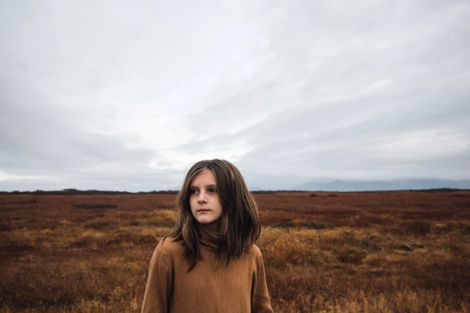 Young girl in a brown ribbed turtleneck standing in a vast open field under an overcast sky, photographed by Christa Renee.