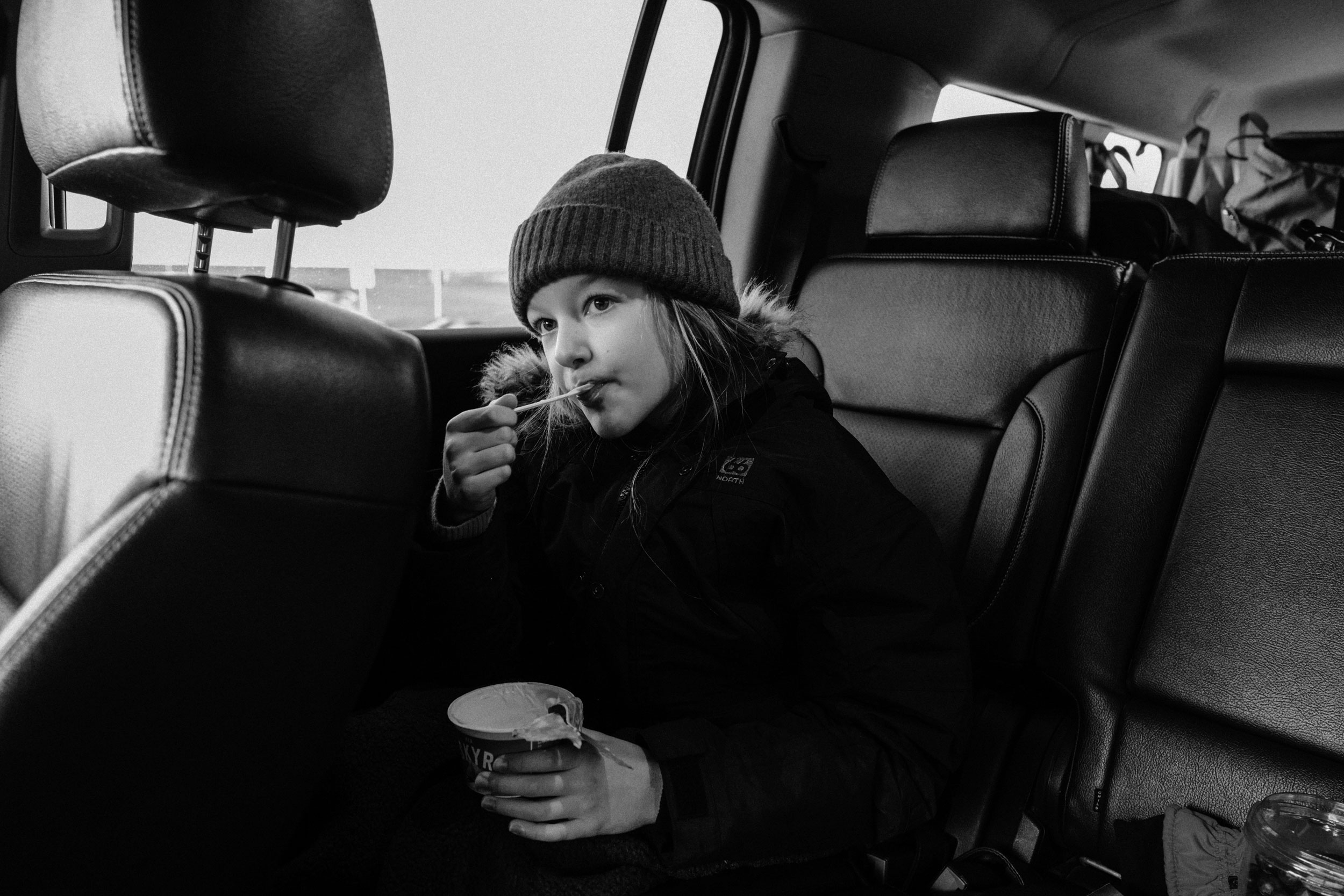 Child wearing a hat and jacket enjoys a cup of yogurt with a spoon while seated in the back of a car on a monochrome image.