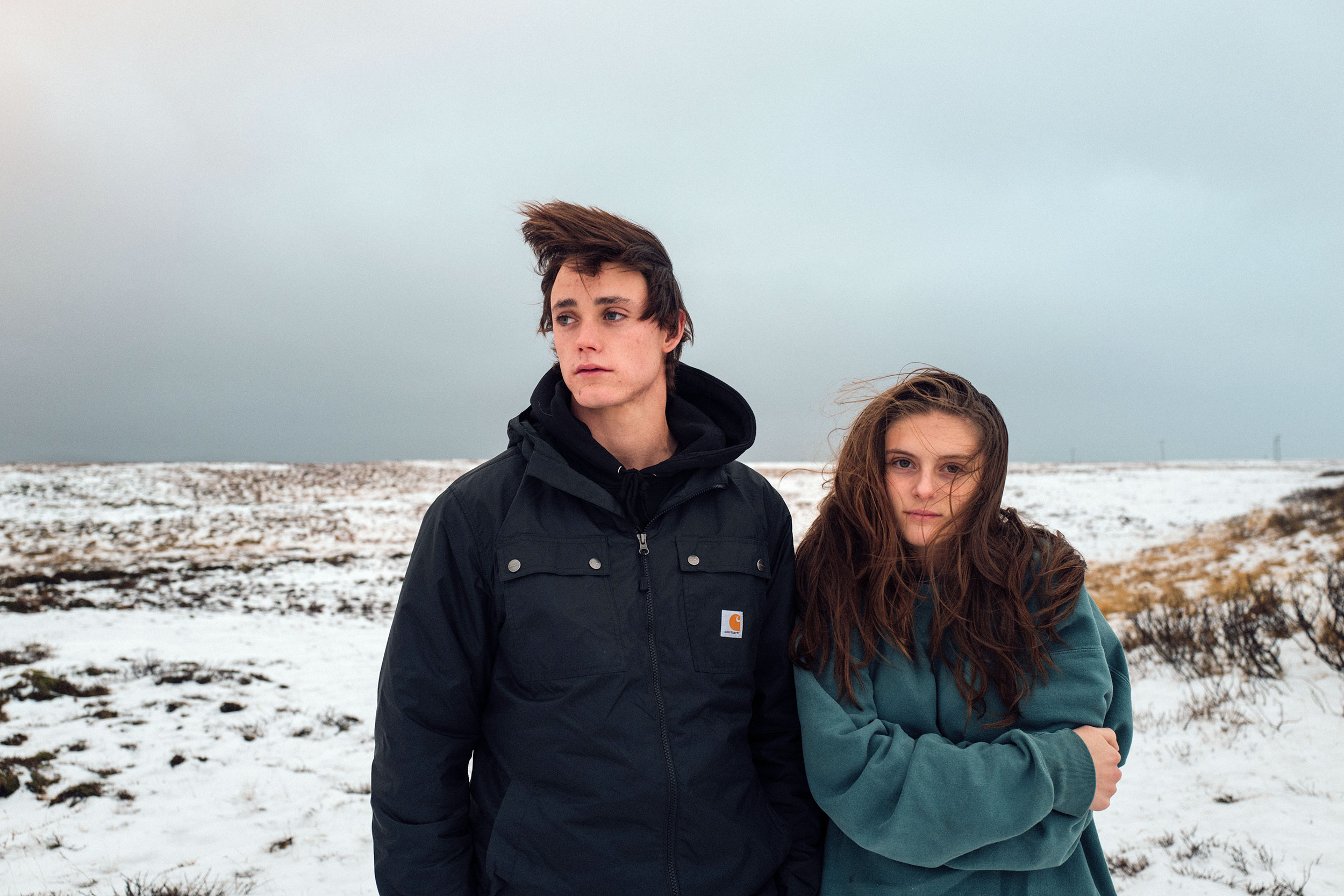 Two young adults, a male and a female, stand outdoors in a snowy field on an overcast day, dressed in jackets. Wind blows through their hair.