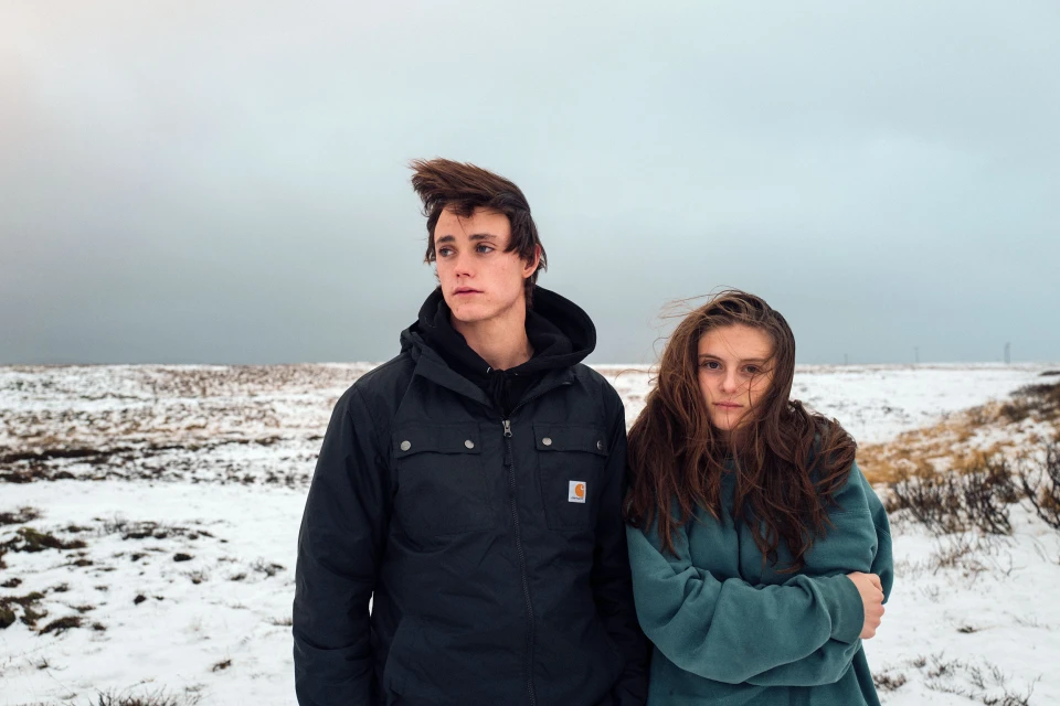 Two young adults, a male and a female, stand outdoors in a snowy field on an overcast day, dressed in jackets. Wind blows through their hair.