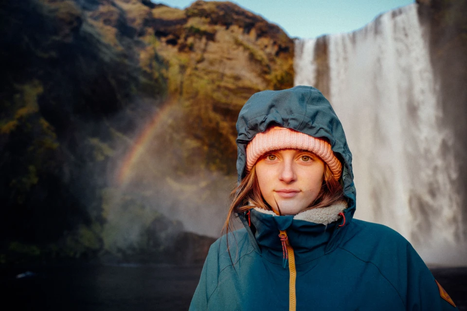 Portrait of a woman wearing a blue hooded jacket and pink beanie, with a waterfall and mountain in the background on a sunny day.