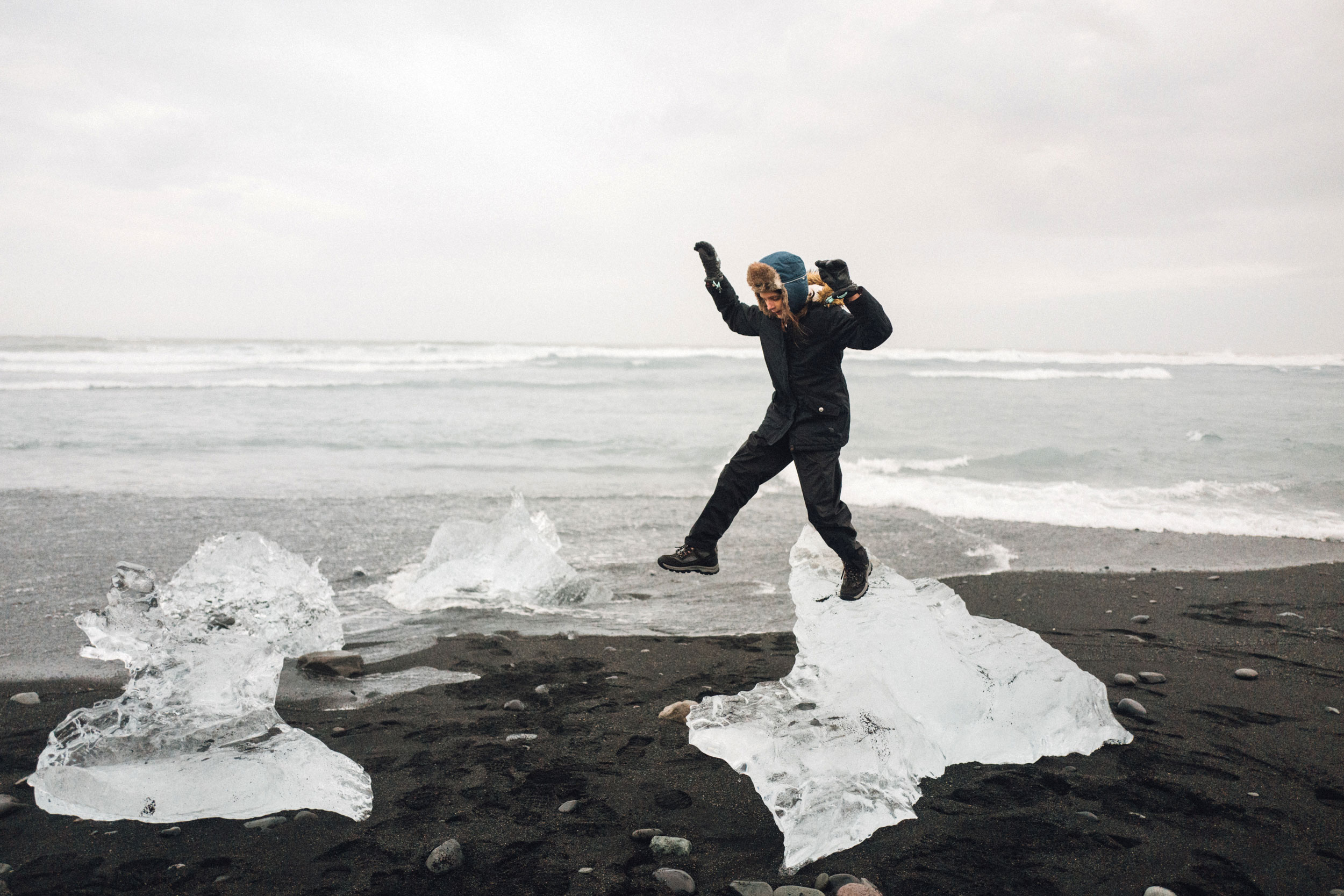 A person jumps between ice formations on a black sand beach near the ocean under a cloudy sky in an outdoor travel scene.