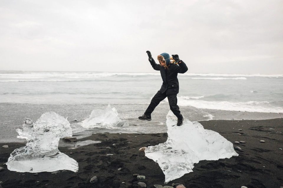 A person jumps between ice formations on a black sand beach near the ocean under a cloudy sky in an outdoor travel scene.