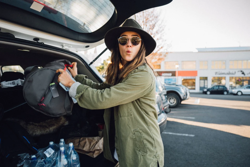 Woman with sunglasses loading a gray duffel bag into the open trunk of a white SUV in a parking lot on a sunny day.