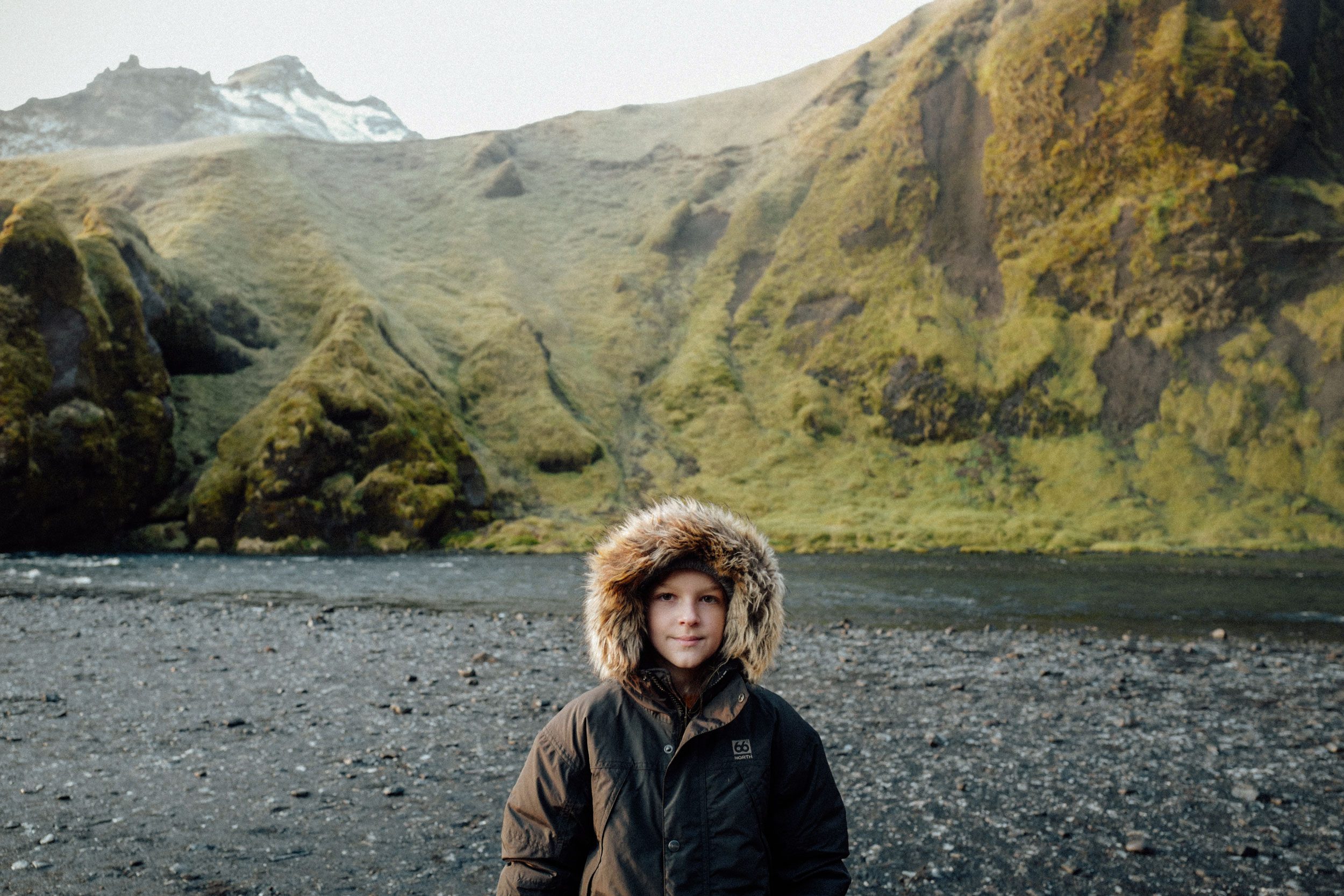 A child in a fur-lined hooded jacket stands in a rocky landscape with a river, with grassy mountains in the background under an overcast sky.