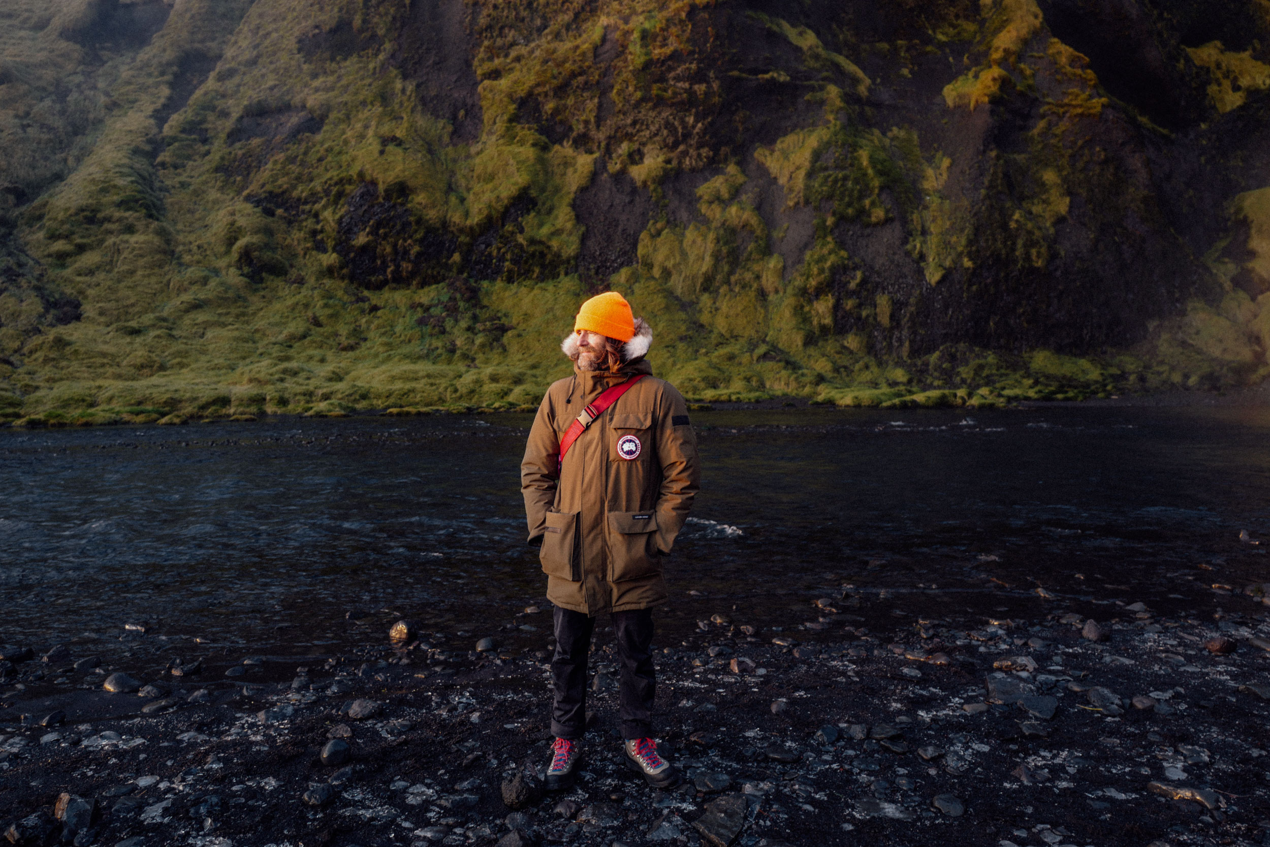 Man stands in front of a black rocky beach with a jacket, orange hat, and a green mossy mountain in the background on a cloudy day.