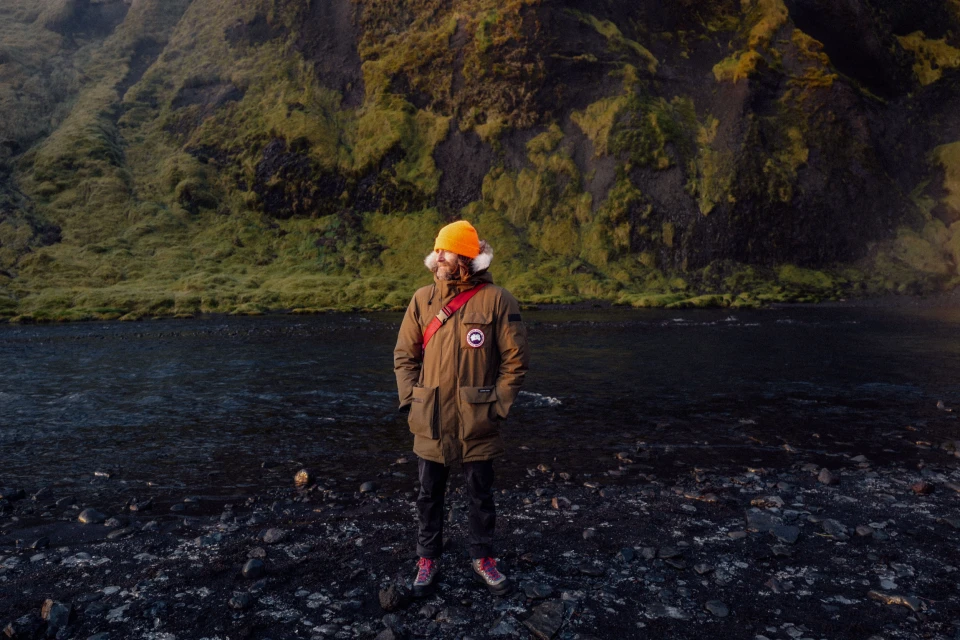 Man stands in front of a black rocky beach with a jacket, orange hat, and a green mossy mountain in the background on a cloudy day.