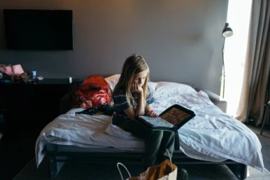 Young child seated on a rumpled bed holding a black binder case inside a contemporary bedroom interior